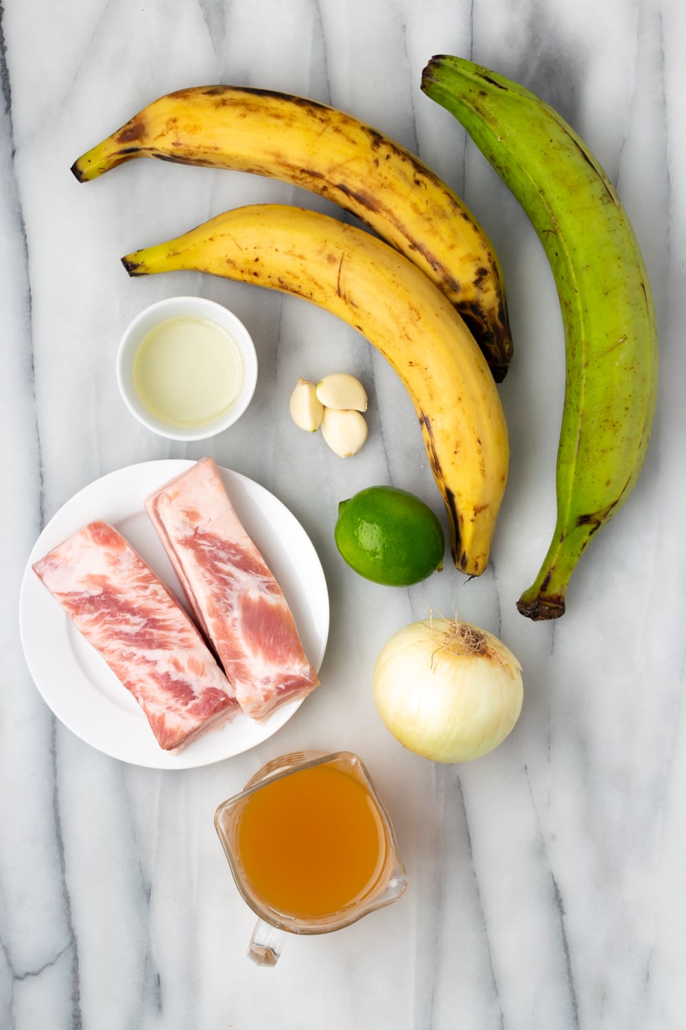 Ingredients for fufu de platano spread on a marble counter: Yellow Plantains, Green Plantain, Olive Oil, Pork Belly, Yellow Onions, Garlic, Lime, Chicken Stock, Salt and Pepper.