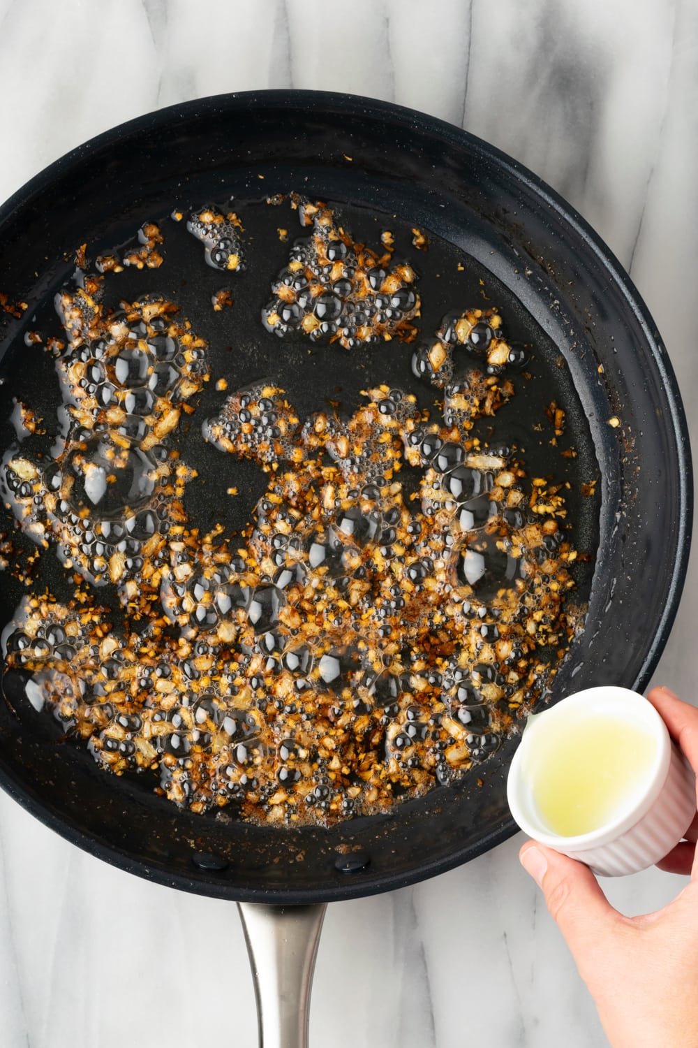 A hand adding lime juice from a small white bowl to a skillet with bubbling pork dripping and cooked onions and garlic on a marble counter.