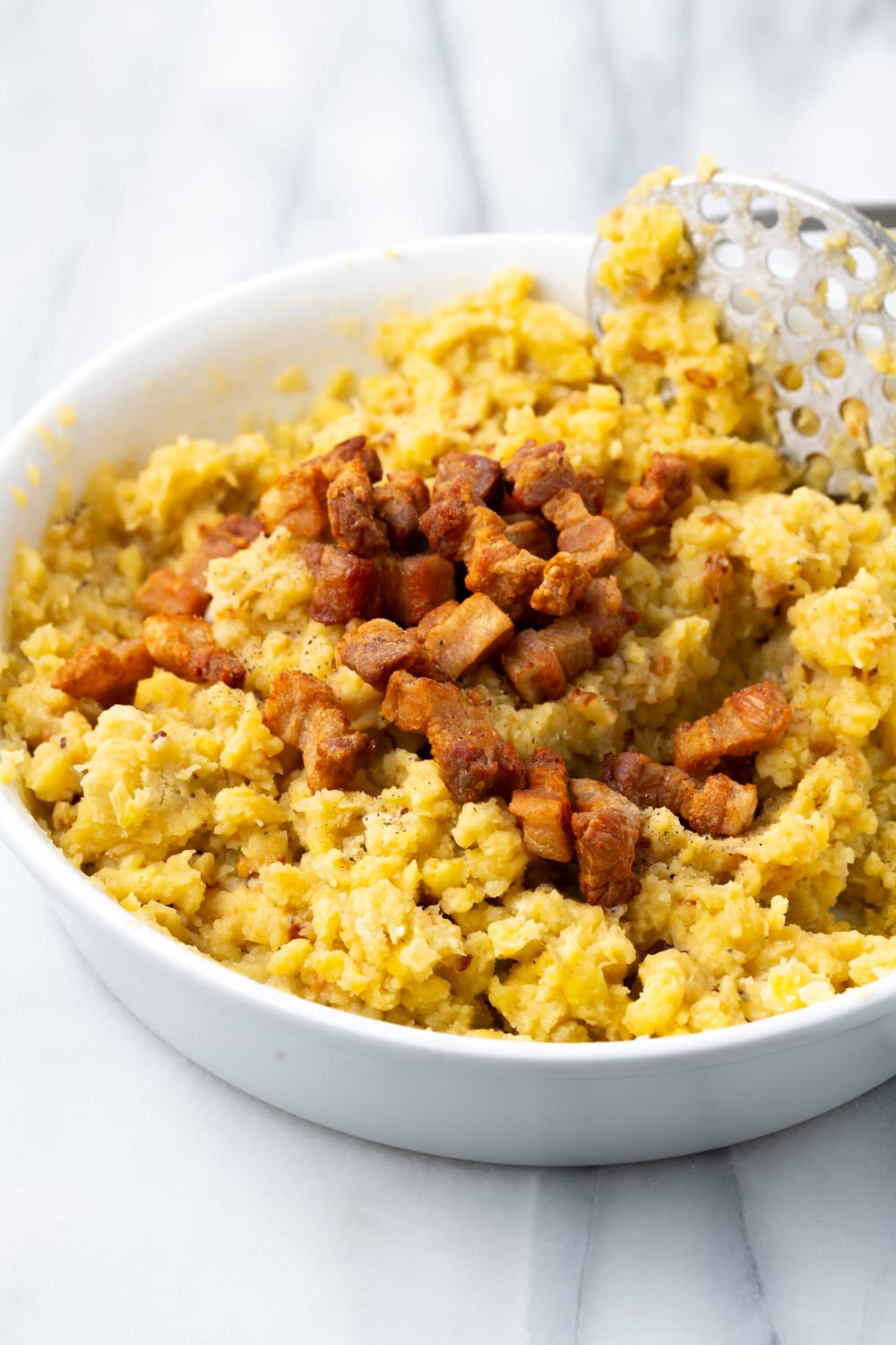 Crispy Pork Belly Pieces being added to mashed plantains in a large white bowl to make fufu de platano with a potato masher in the bowl too.